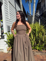 Woman in a brown dress standing outdoors with palm trees and a building in the background